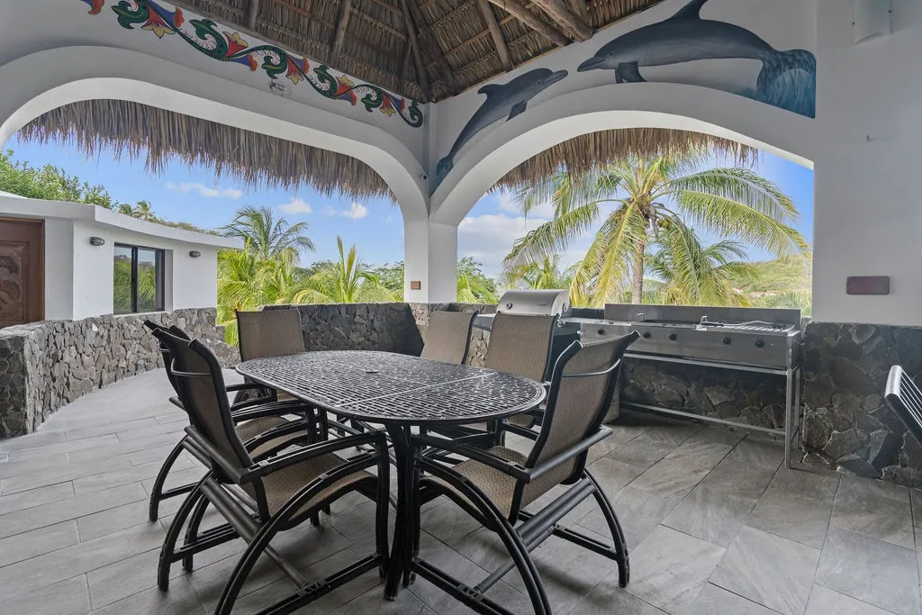 Outdoor dining table under the thatched rancho with dolphin murals, grill, and palm views
