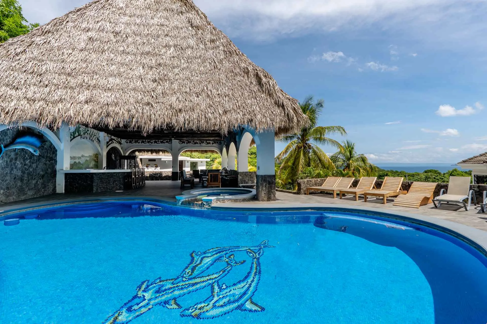 Pool with dolphin mosaic and waterfall jacuzzi beside the thatched rancho, loungers and the Pacific beyond