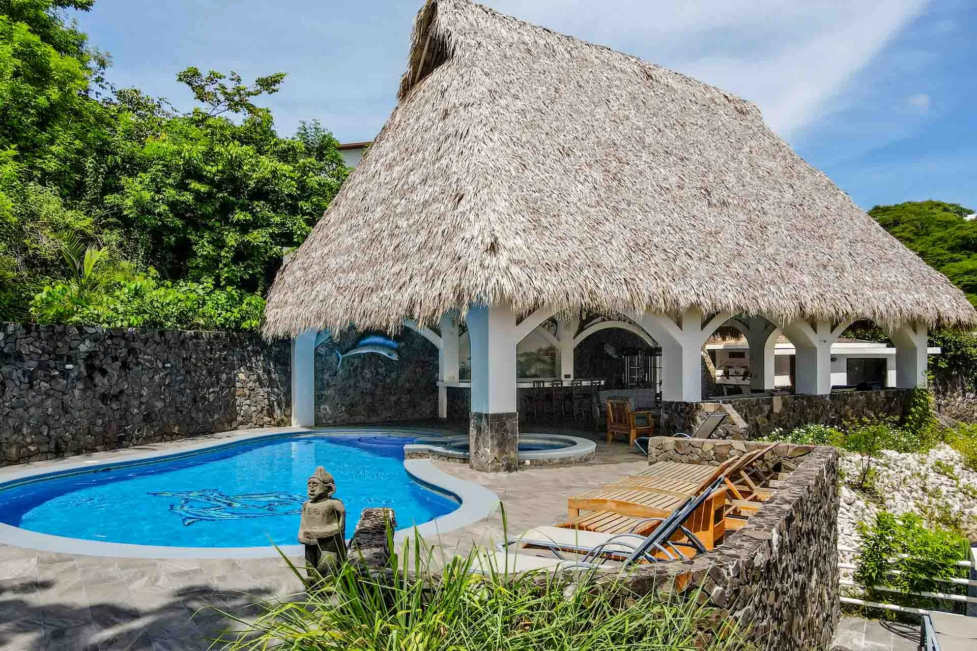 Thatched rancho and pool seen from the garden, with stone walls and wood loungers